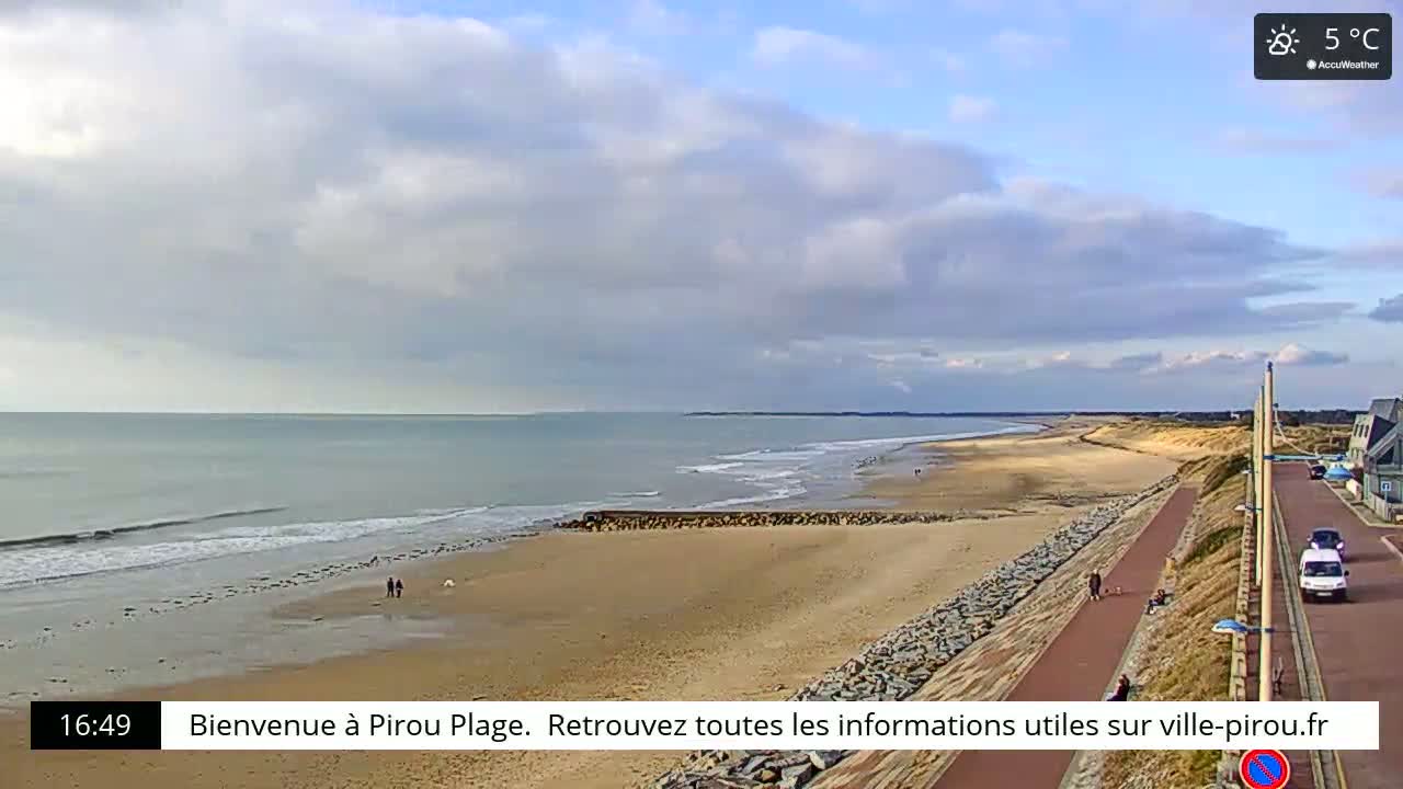 A long sandy beach stretches along a calm ocean under a partly cloudy sky, with a paved walkway and a few people visible.