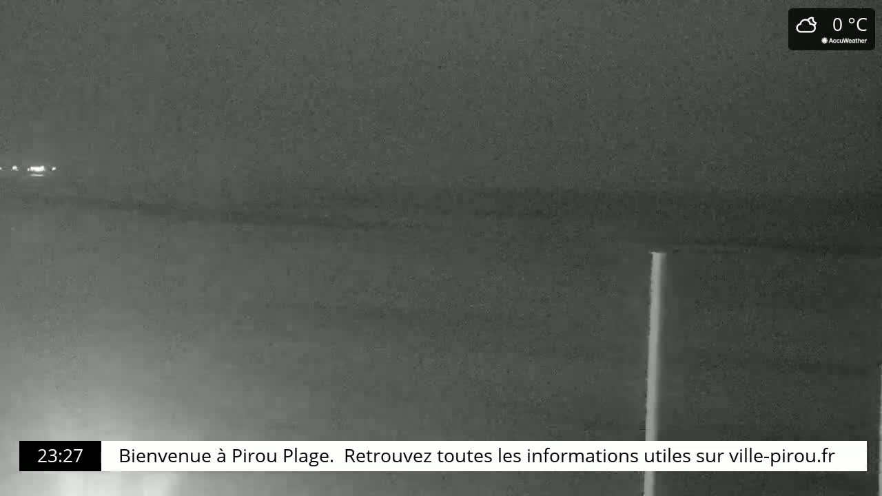 A dark, nighttime view of a beach with a single, bright vertical pole and faint lights in the distance at 0°C.