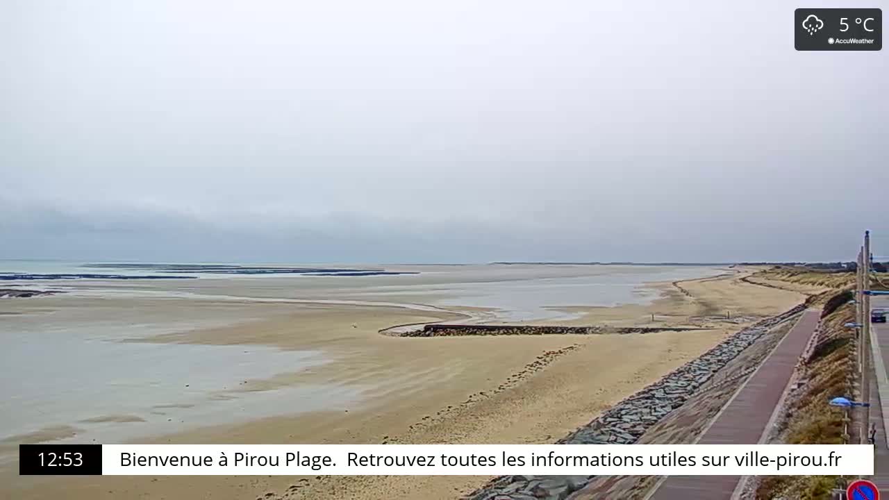 A wide sandy beach stretches to a calm, light-grey ocean under an overcast sky, with a paved walkway along the shore on a 5°C day.