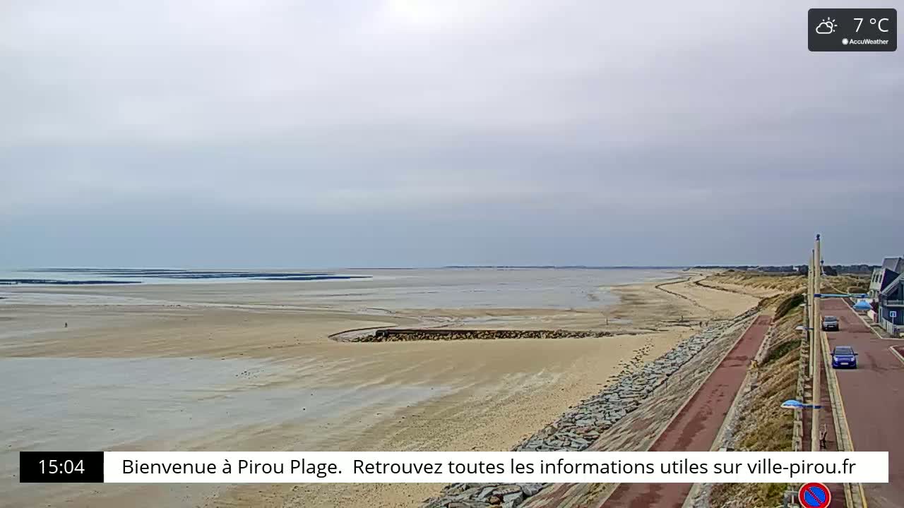 A mostly cloudy day shows a wide sandy beach next to a calm ocean, with a paved road and a few cars visible in the upper right corner.