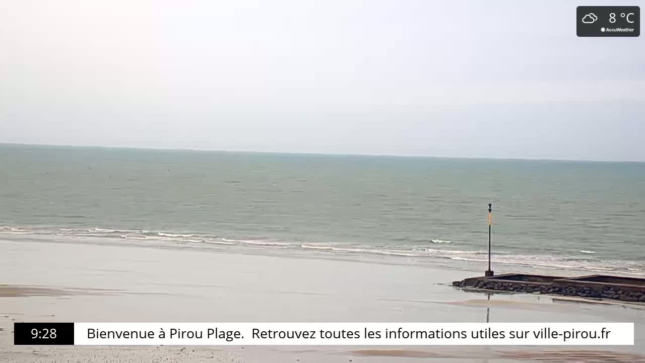 A mostly cloudy day shows a calm ocean meeting a wide sandy beach with a small structure at the shoreline.