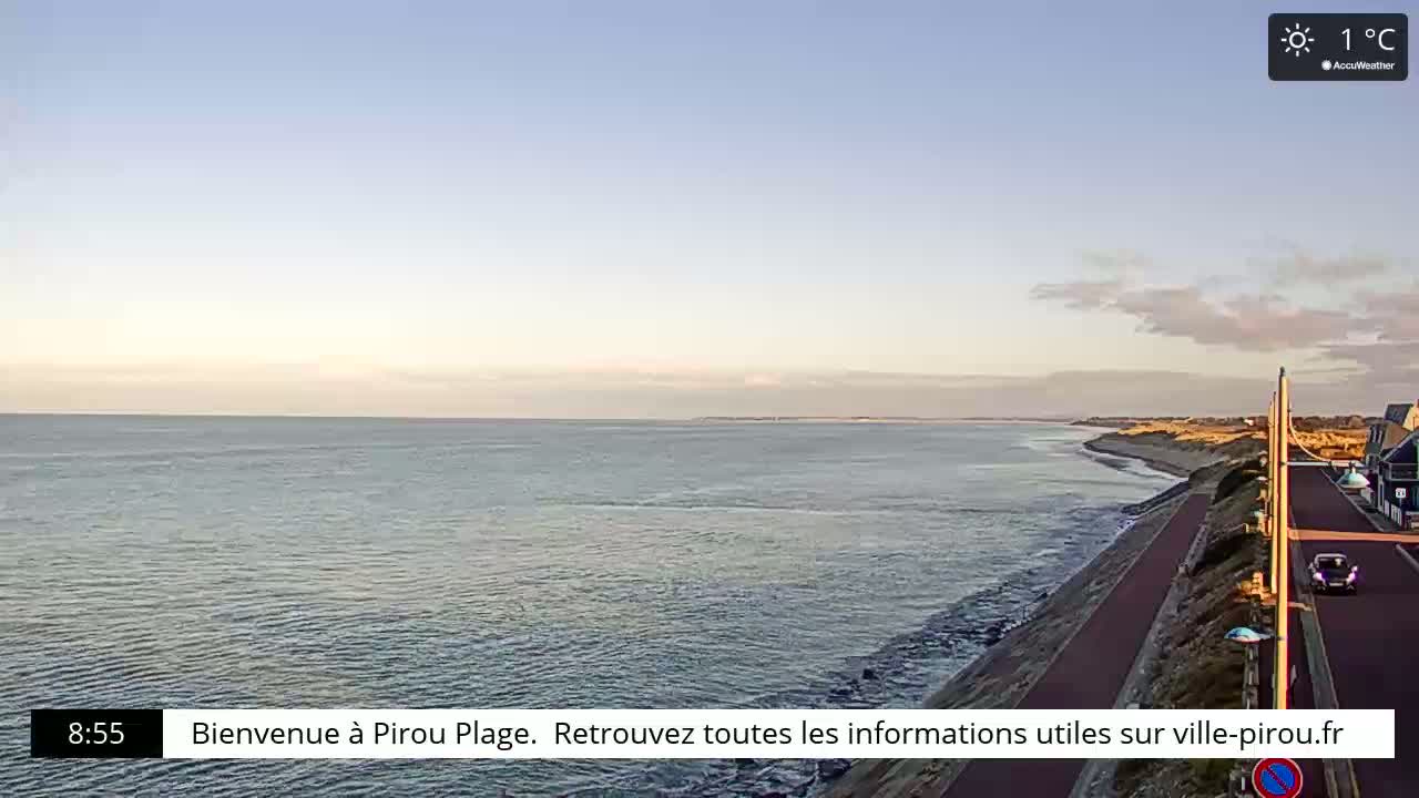 A calm ocean meets a sandy shoreline with a paved path and sparse buildings under a mostly clear sky, with a temperature of 1°C.