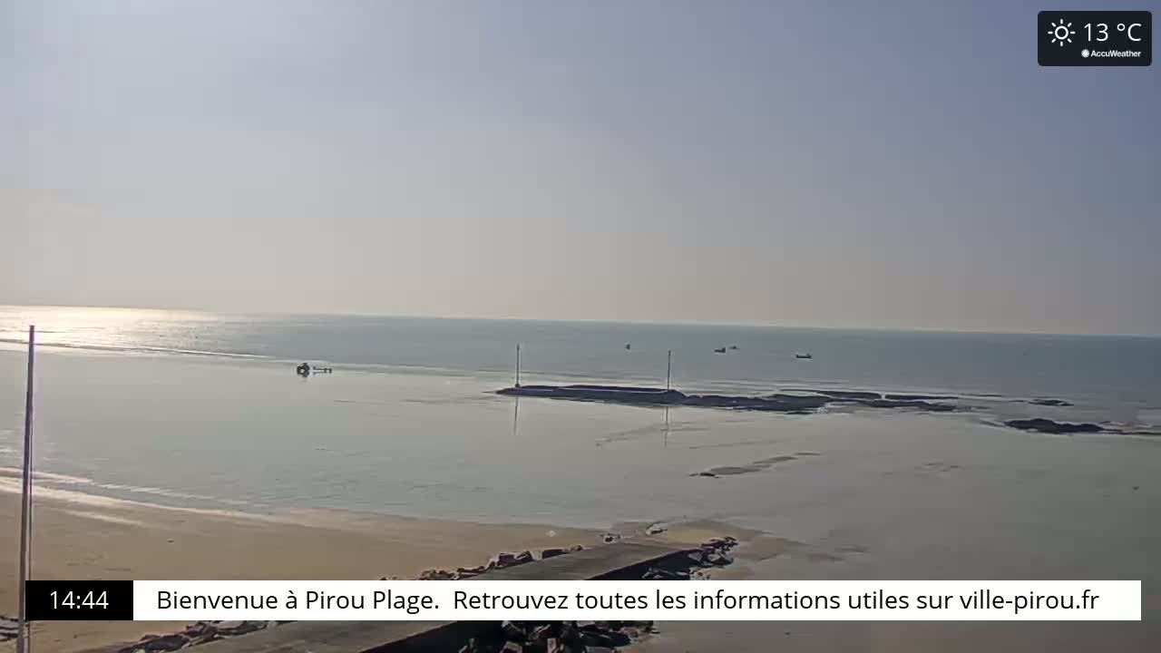 A calm, sunny beach scene shows a sandy shore meeting a tranquil body of water under a clear, pale sky.