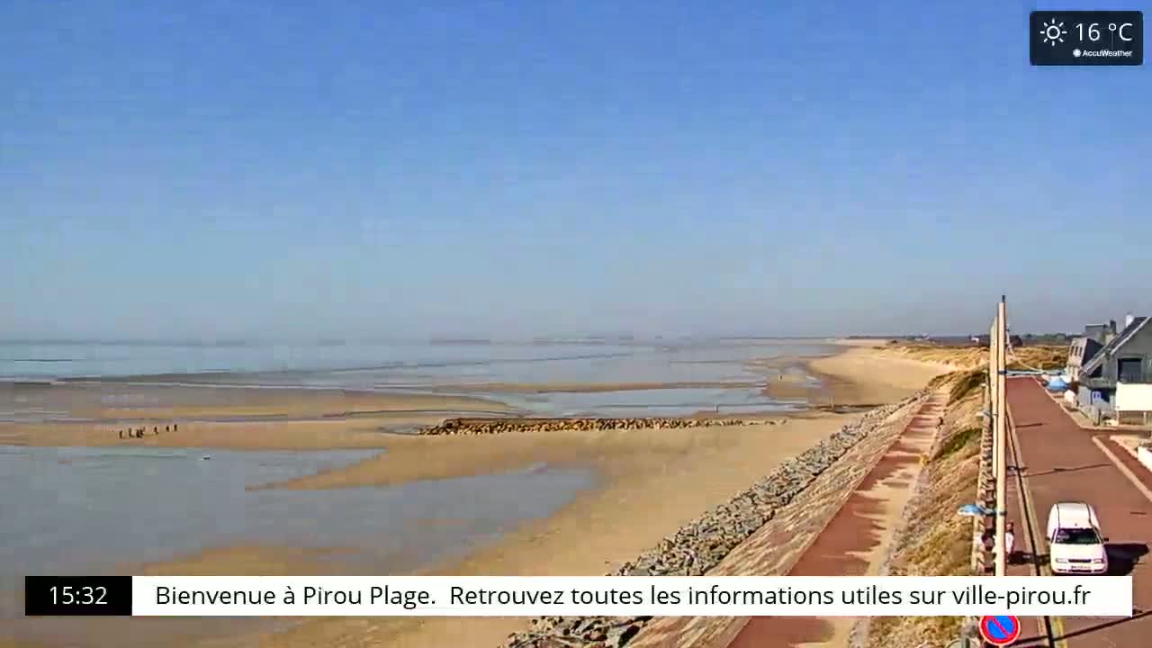 A wide sandy beach stretches along a calm, shallow ocean under a clear blue sky, with a paved walkway and buildings visible in the background.