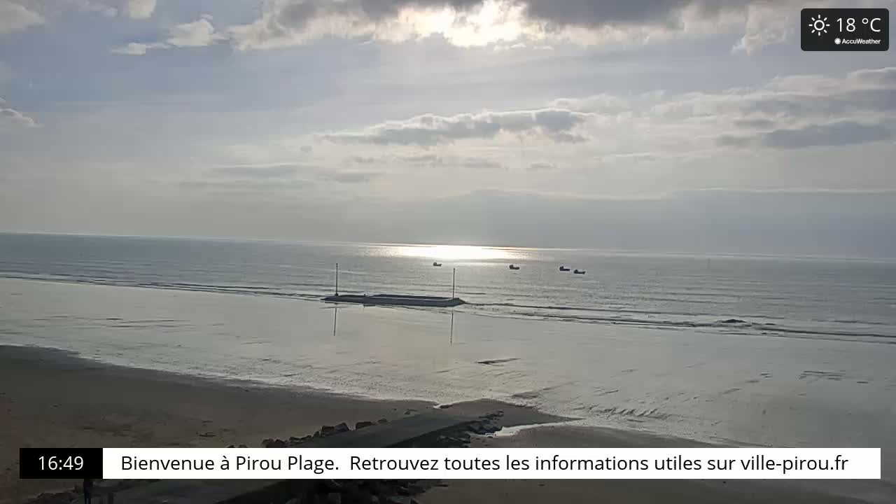 A partly cloudy, sunny day at a beach with calm water and several small boats visible in the distance.