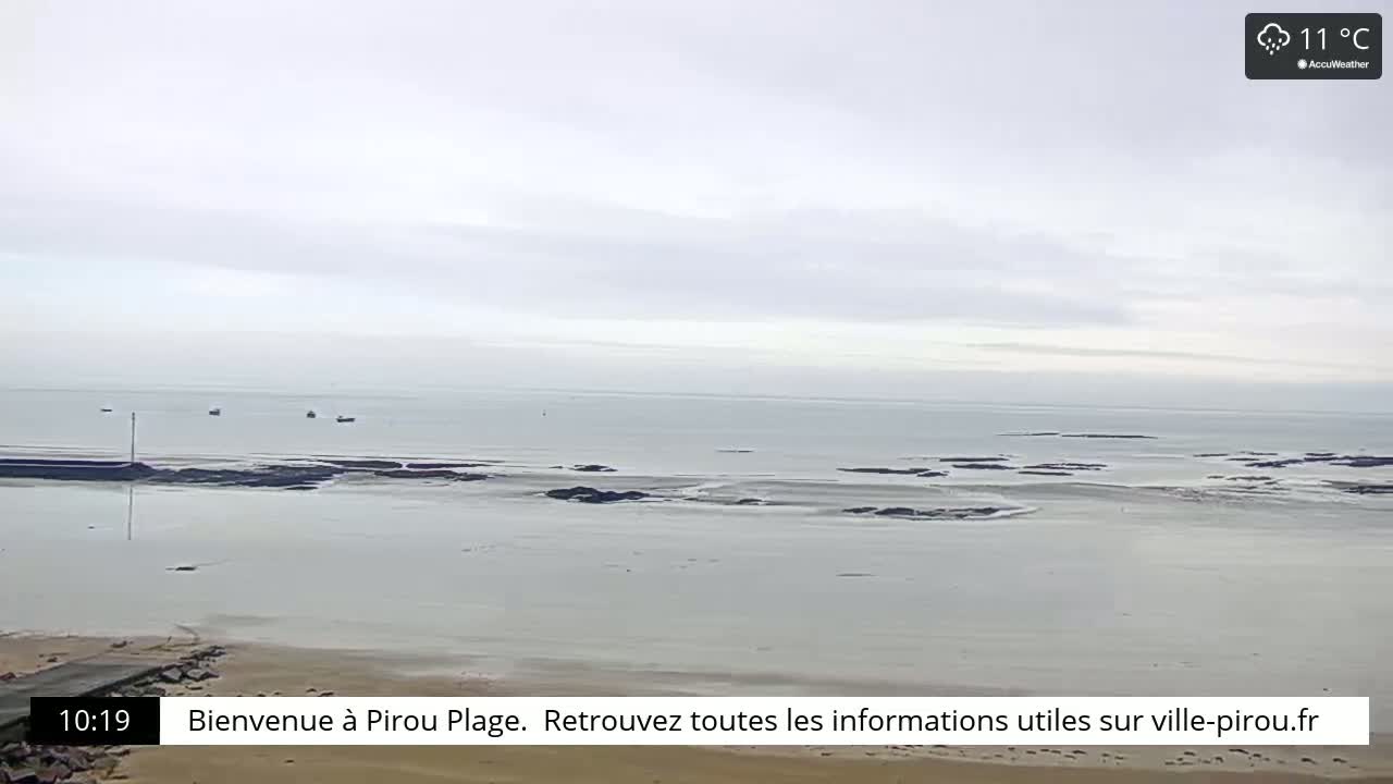 A calm, overcast day at a beach shows a mostly flat, light gray ocean with some dark rocks visible near the shore, a few small boats in the distance, and a sandy beach.