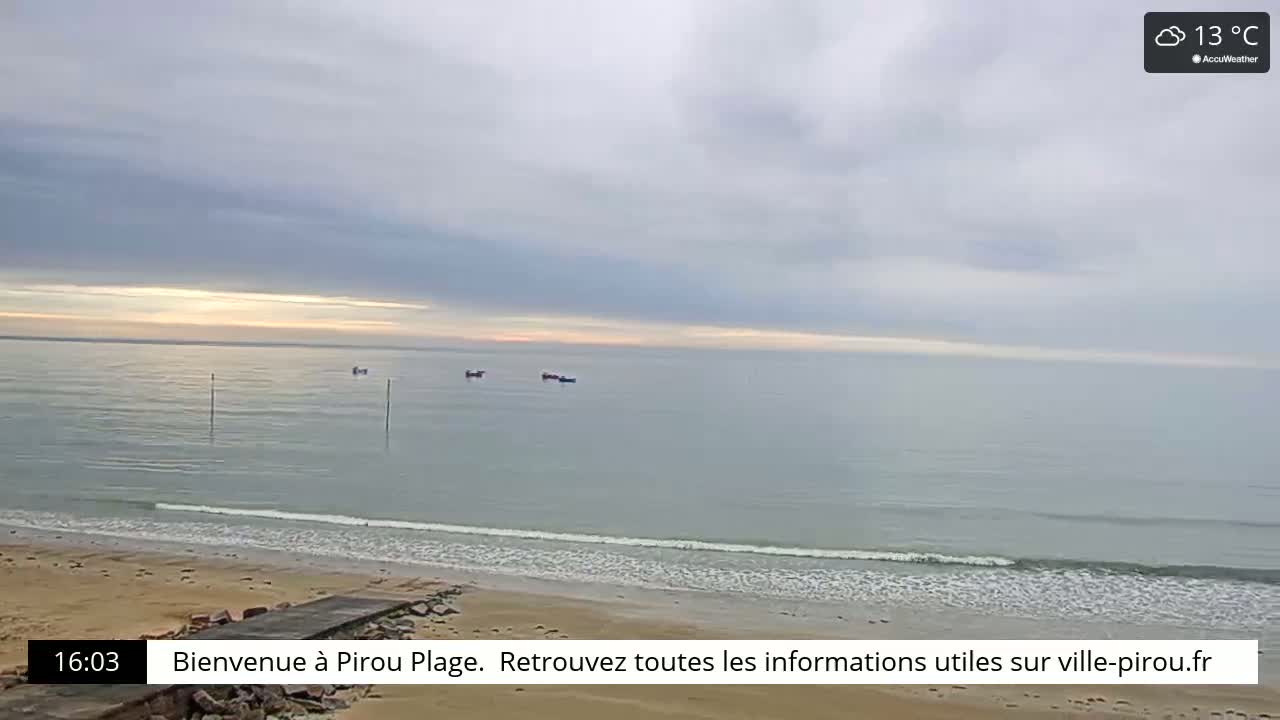 A sandy beach meets a calm, grey ocean under an overcast sky, with a few small boats visible on the water.