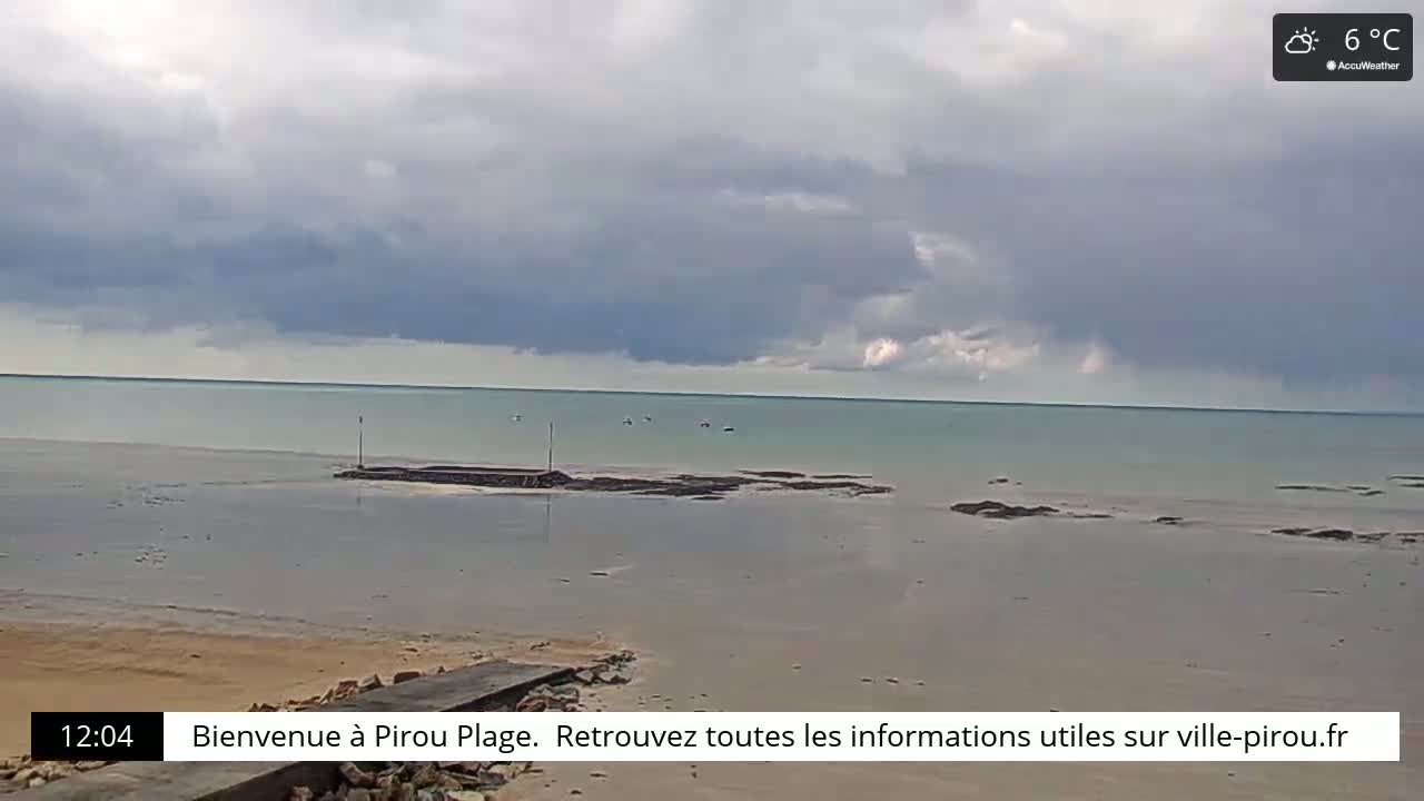 A mostly cloudy, cool day at a beach with calm, light-green water, showing a sandy shore and a low-lying stone structure extending into the water.