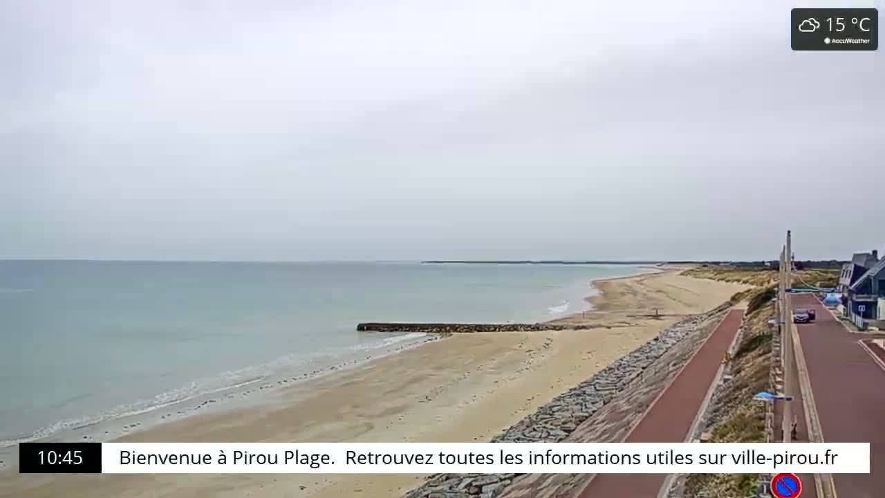 A sandy beach with calm, light-blue water stretches out under an overcast sky, a paved walkway and road running alongside it.