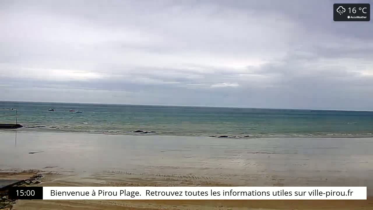 A mostly overcast sky hangs over a calm, teal ocean meeting a wide, sandy beach at low tide, with a few small boats visible on the water.