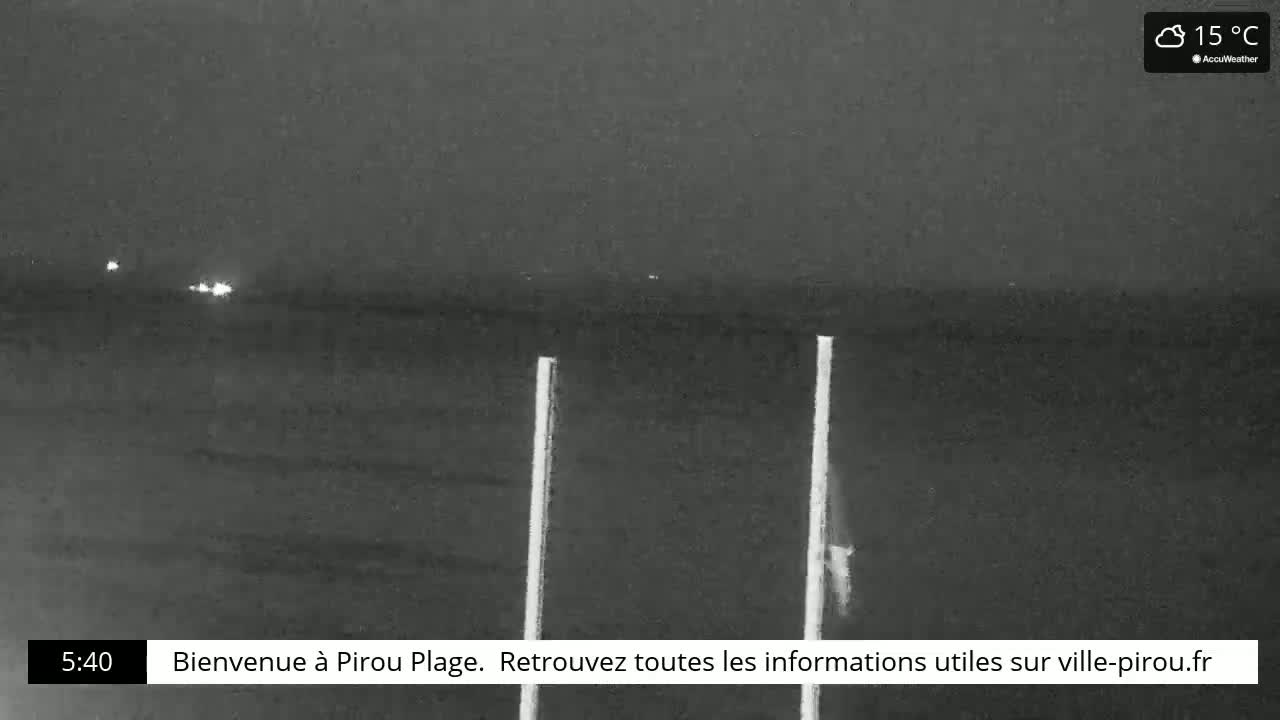 A nighttime, low-light view of a dark beach and ocean under a cloudy sky, with two tall, thin, light-colored poles in the foreground.
