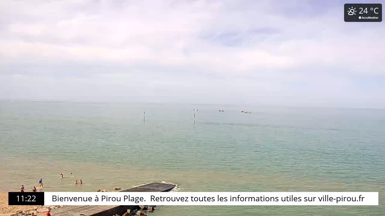 A calm, light-grey day shows a beach with a few people swimming in the shallows, a calm sea extending to the horizon, and several small boats in the distance.