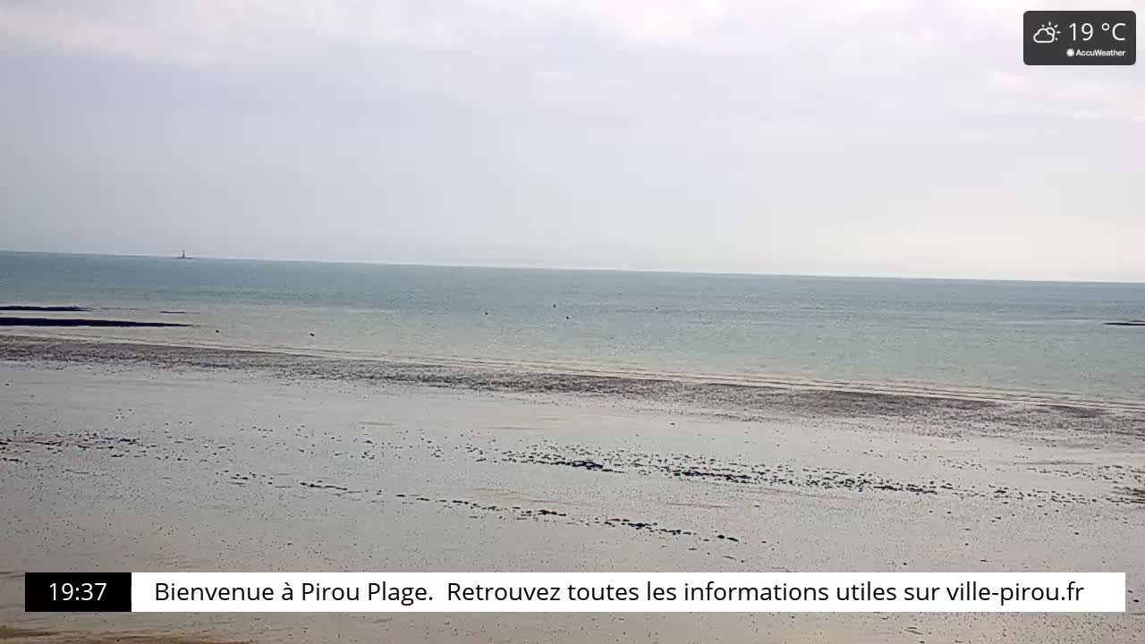 A wide expanse of sandy beach meets a calm, light-blue ocean under an overcast sky, with a distant lighthouse visible on the horizon.