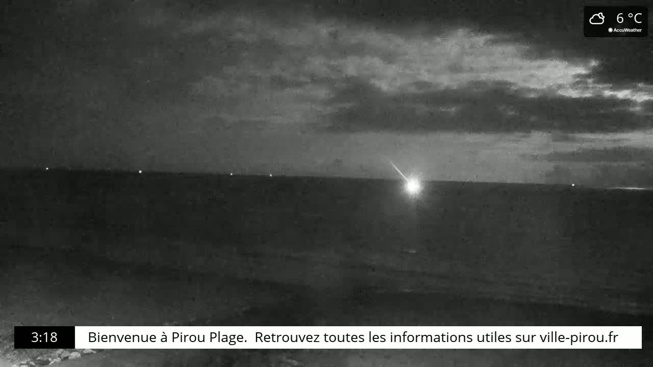 A nighttime, cloudy coastal scene shows a bright meteor streaking across the dark sky over a calm body of water.