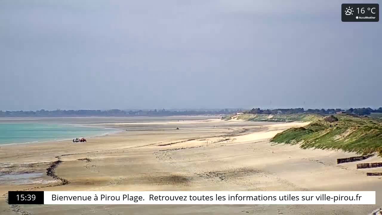 A long sandy beach curves gently into a calm, light-blue ocean under a mostly clear sky, with a few people visible in the distance and green dunes bordering the beach.