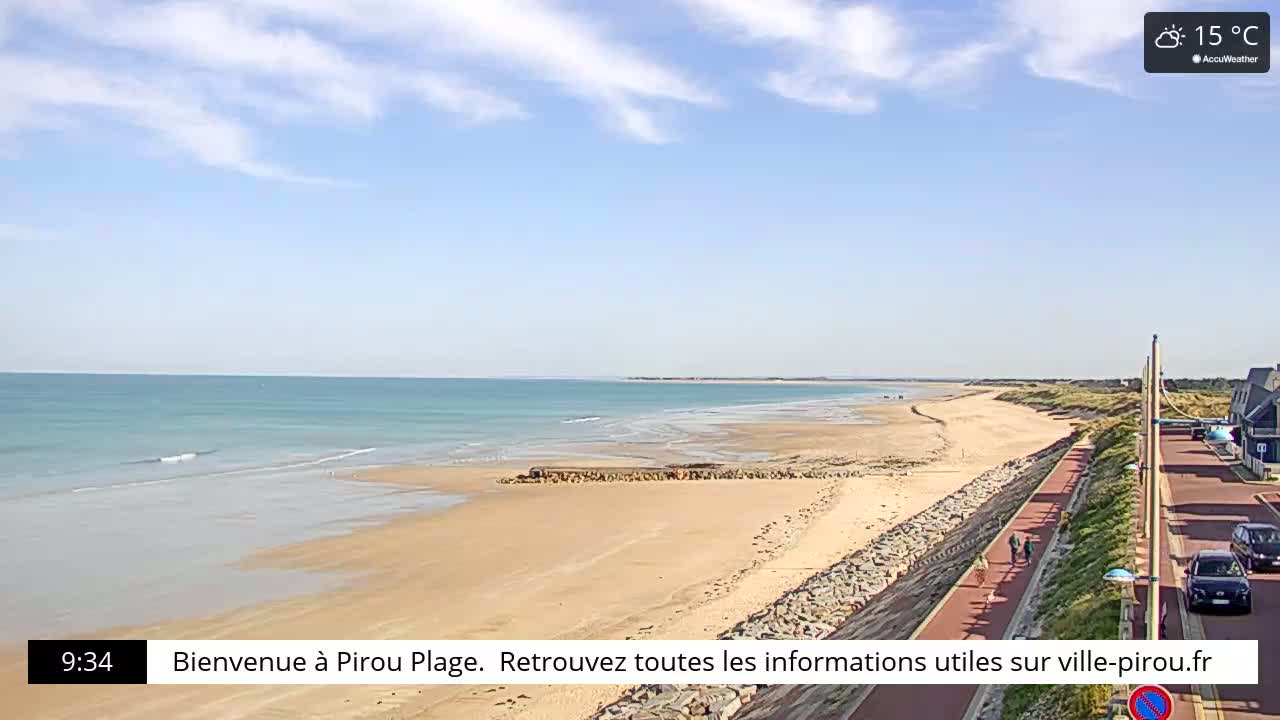 A sandy beach stretches along a calm, light-blue ocean under a mostly clear sky, with a paved walkway and several cars visible nearby.