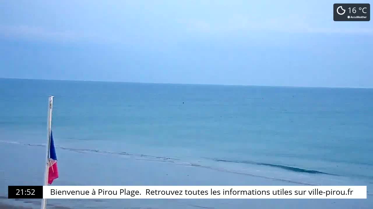 A French flag stands on a beach next to a calm, light blue ocean under a clear, pale blue sky.
