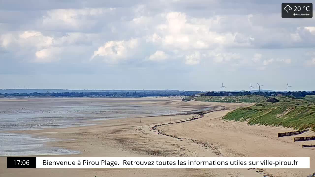 A wide sandy beach curves gently to the left, under a partly cloudy sky, with wind turbines visible in the distance.