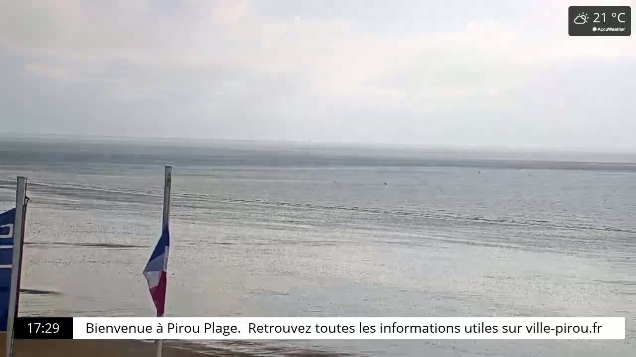 A mostly overcast beach scene shows a calm, wide expanse of water receding to reveal a sandy shoreline, with a French flag displayed on a pole.