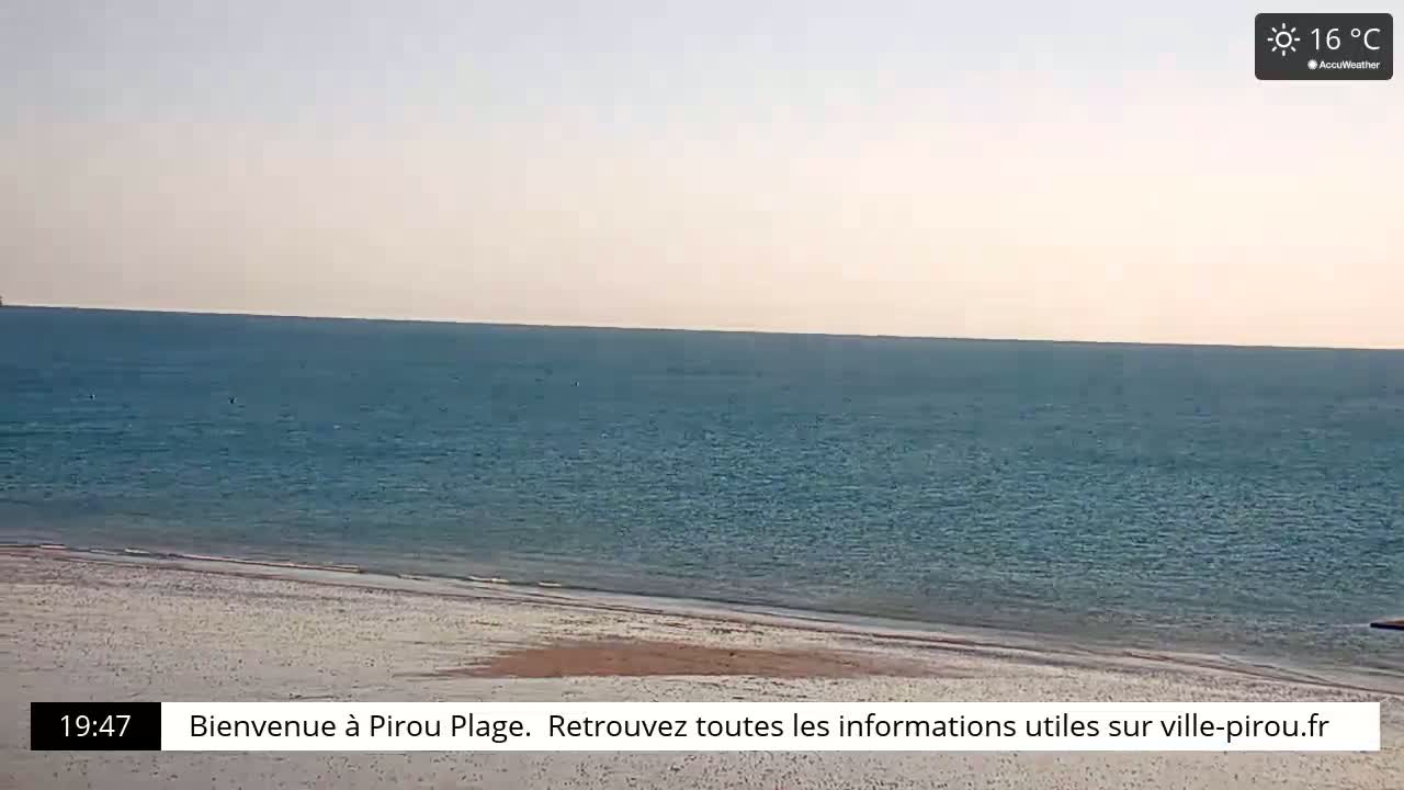 A calm, blue ocean meets a sandy beach under a clear, pale sky on a 16°C day.