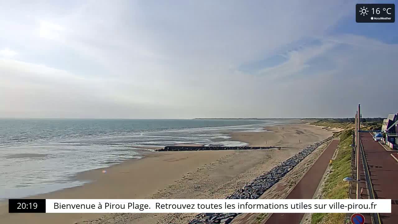 A sandy beach stretches along a calm ocean under a partly cloudy sky, with a paved walkway and dune beside it.