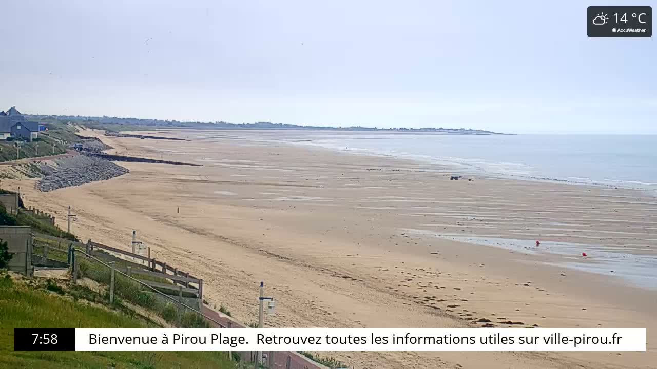 A long, sandy beach stretches to a calm ocean under a clear sky, with a coastal path and buildings visible in the distance.