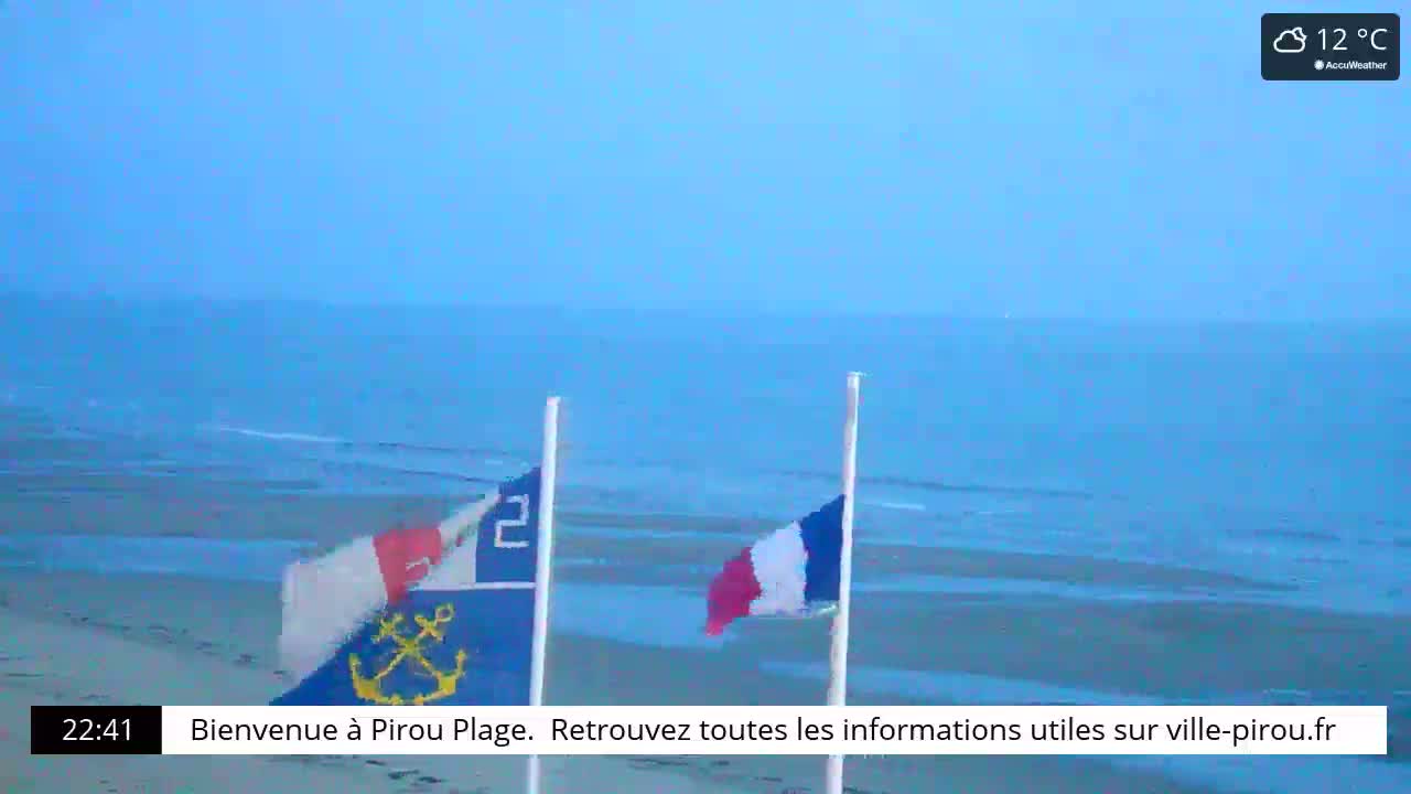 Two flags wave on a beach next to a calm sea under a cloudy sky.