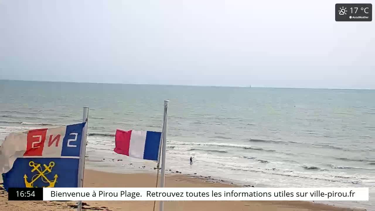 A sandy beach with calm ocean waves under a mostly clear sky displays two flags, one with nautical symbols, the other the French tricolor.