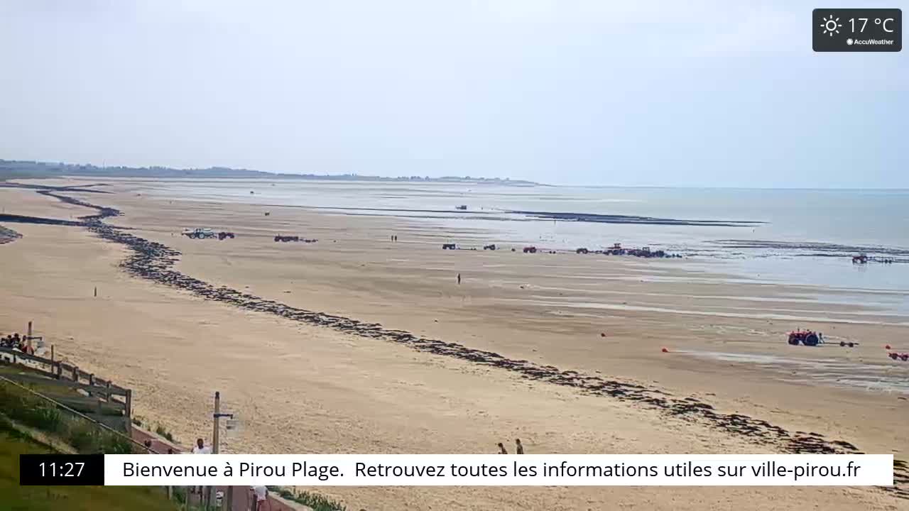 A long sandy beach stretches to a calm ocean under a clear sky, with several vehicles and a few people visible on the sand.