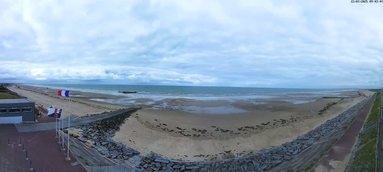 An aerial view shows a wide sandy beach, a calm sea under a cloudy sky, and a stone seawall with a paved path beside it.