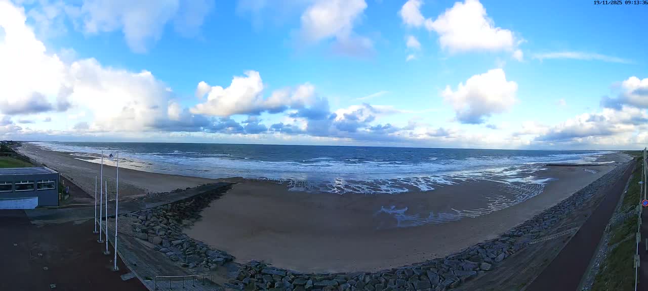 A wide panoramic view captures a sandy beach and a choppy ocean under a partly cloudy sky, with a rocky seawall and paved path running alongside the shore.