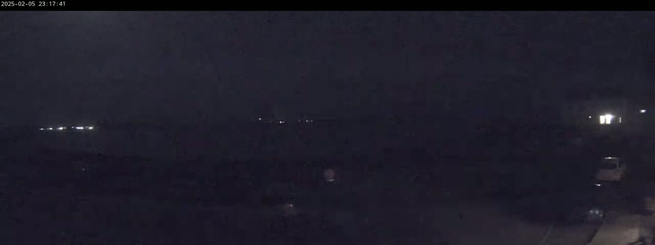 A nighttime, dark scene shows a distant line of headlights and a few illuminated windows in the far right, with a dark, mostly featureless foreground.