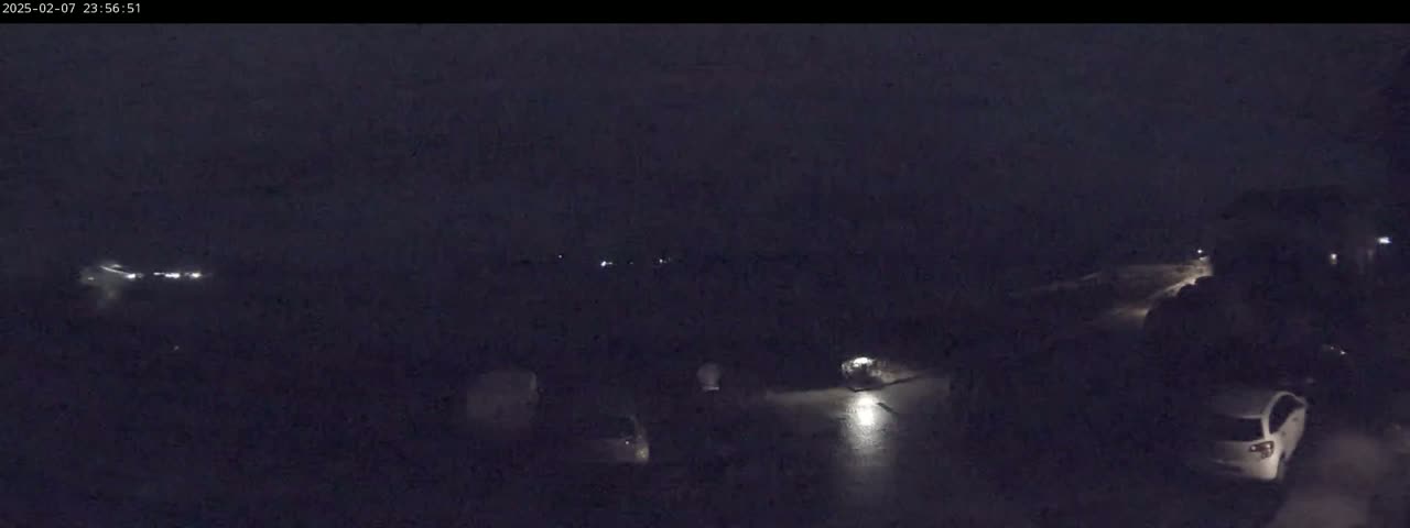 A dark, nighttime scene shows several vehicles parked or stopped in a lot, with some faint lights visible in the distance.