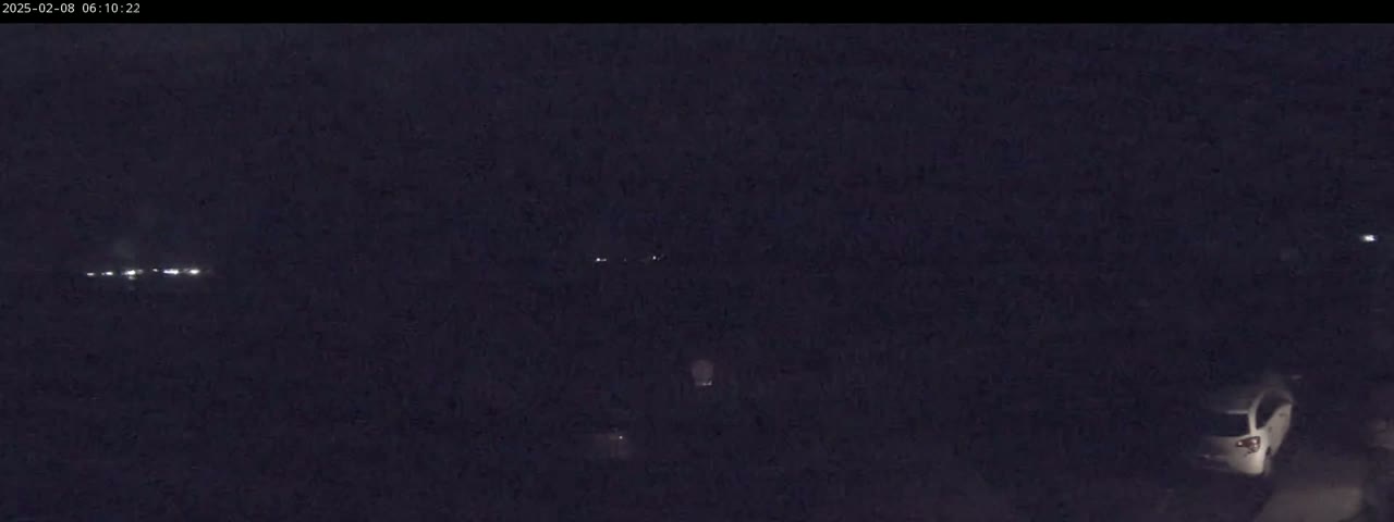 A dark, nighttime scene shows a few distant lights and a single light-colored vehicle in the lower right corner.