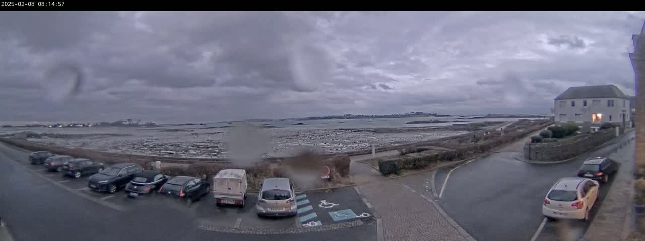 A parking lot with several cars is situated next to a road overlooking a calm body of water under an overcast sky.