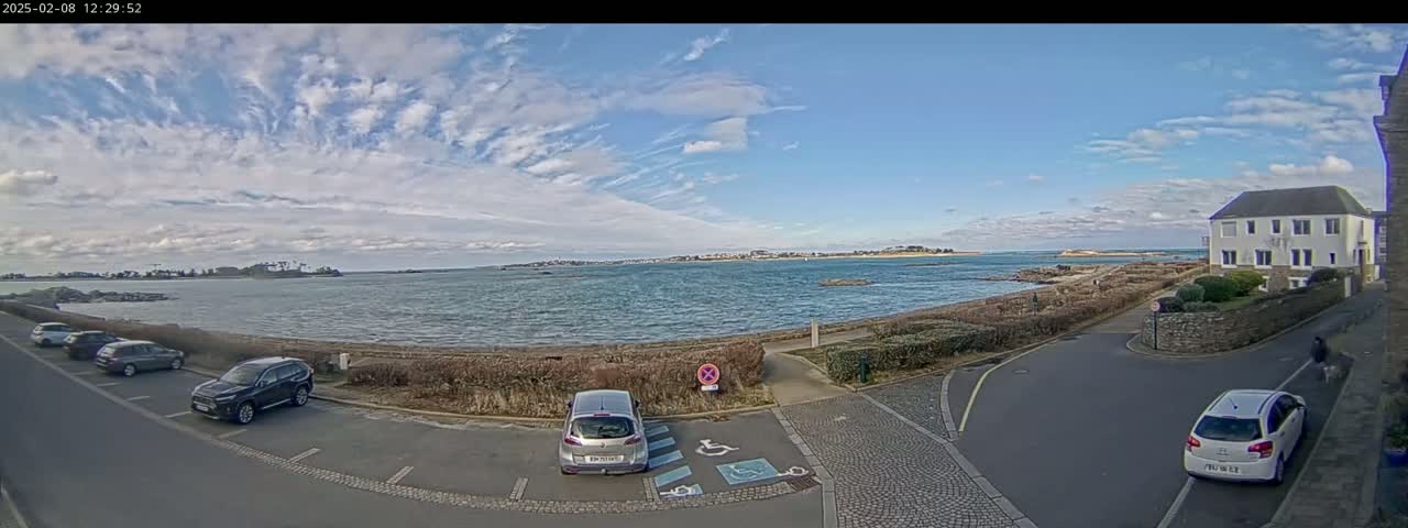 A partly cloudy sky overlooks a calm body of water with a few cars parked near a residential building.
