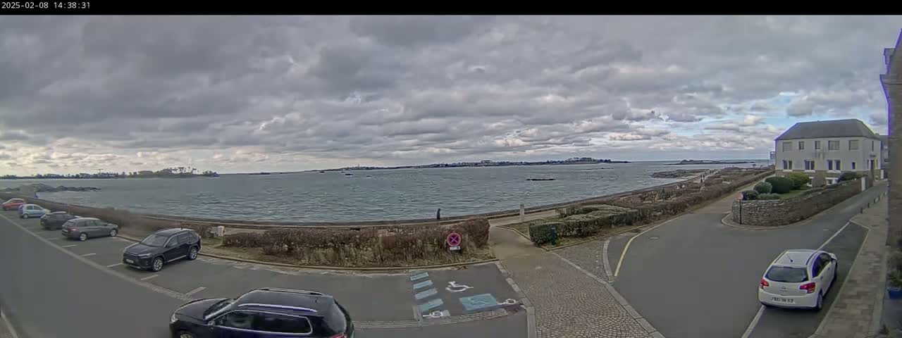 A parking lot overlooks a choppy sea under a cloudy sky, with several cars parked and a building visible in the distance.