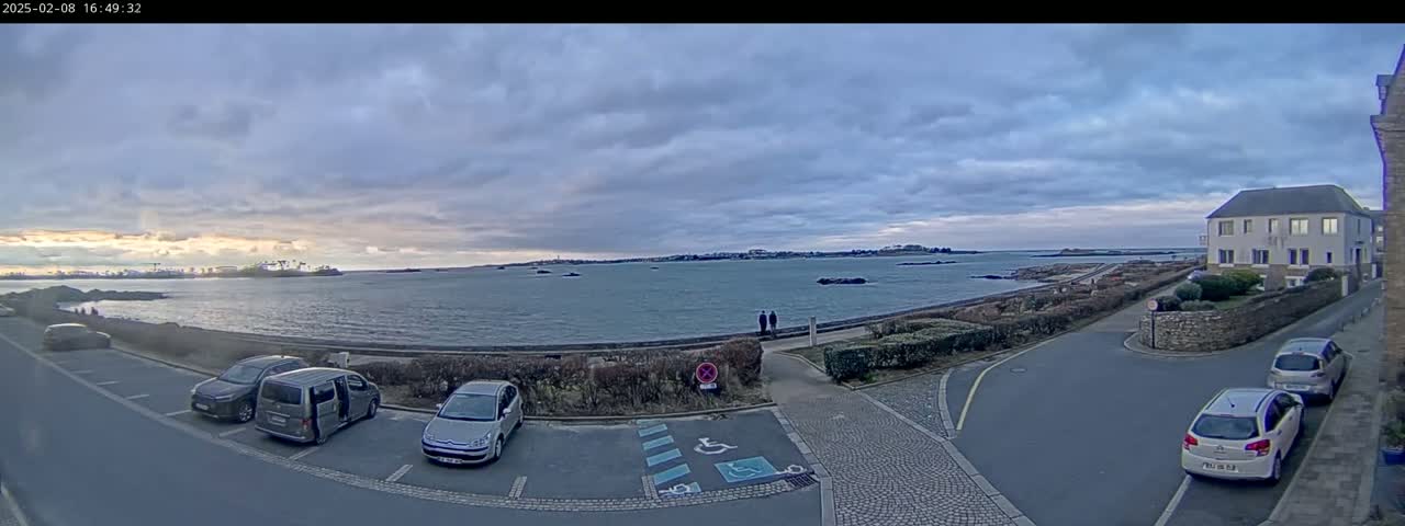 A seaside parking lot with several cars is situated near a waterfront path and a residential building under a cloudy sky.