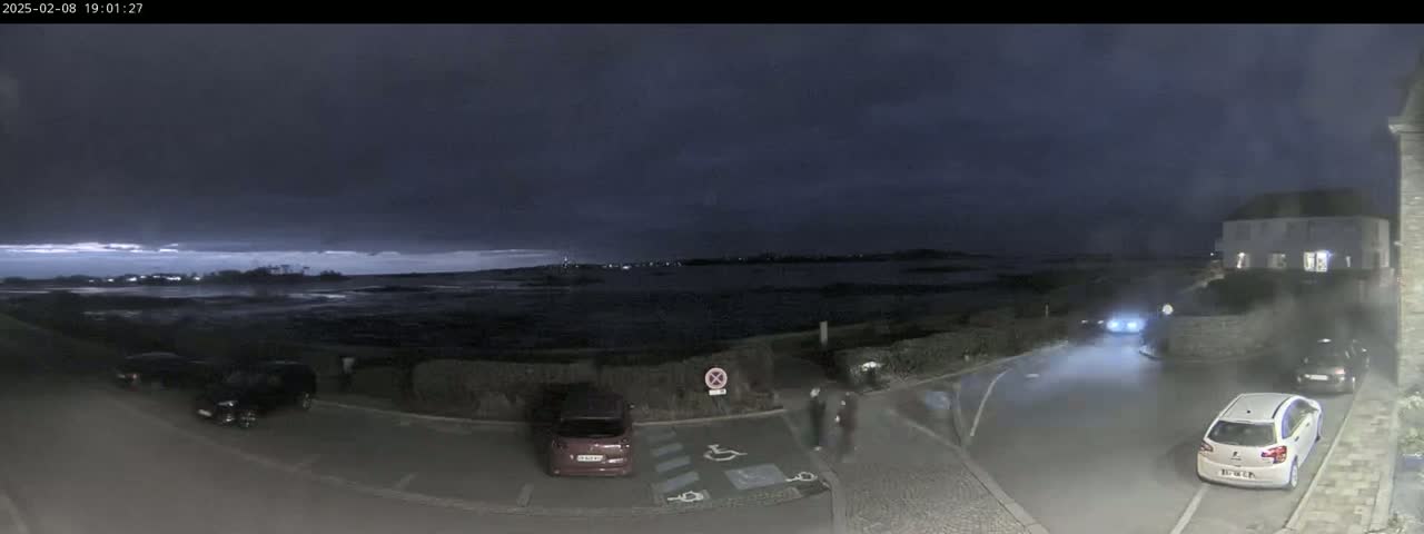 A dark, overcast night scene shows a parking lot with several cars near a body of water, with a few people visible near the edge of the lot.