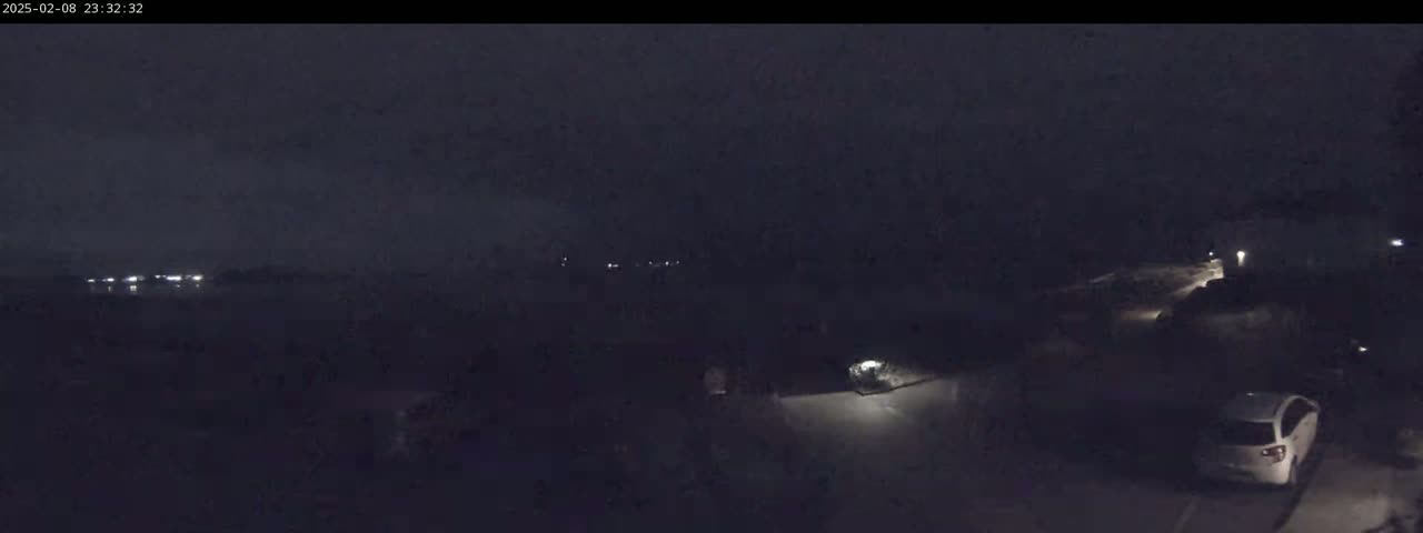 A nighttime, low-light view shows a dark, possibly overcast sky above a sparsely lit area with a few vehicles visible, including one parked car.