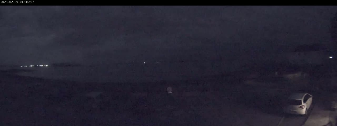 A dark, nighttime scene shows a few distant lights and a white car parked in a dimly lit area under an overcast sky.