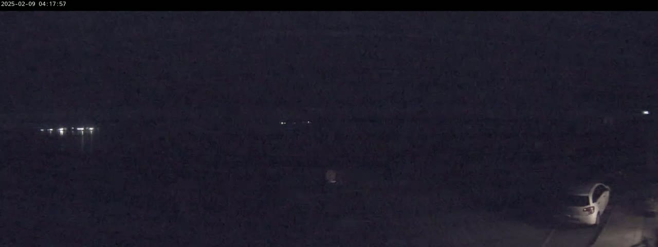 A dark, nighttime scene shows a car parked near a body of water with distant lights visible across the water.
