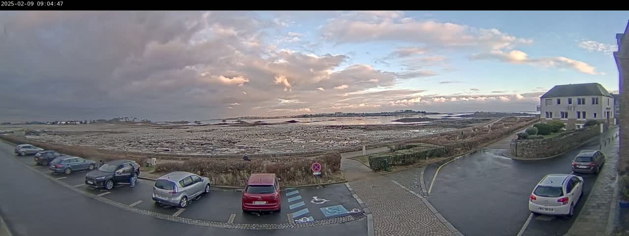 A parking lot with several cars is next to a low stone wall overlooking a wide expanse of tidal flats and calm water under a partly cloudy sky.