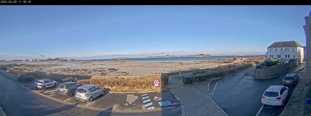 A clear, sunny day reveals a coastal scene with several cars parked near a building, overlooking a rocky shore and calm water extending to a distant landmass.