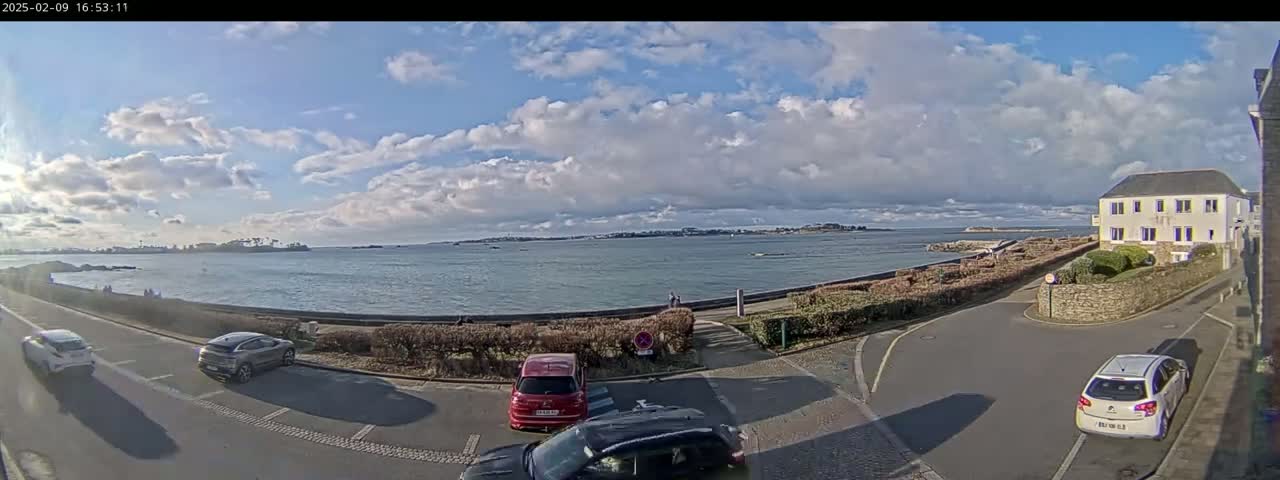 A partly cloudy day overlooks a calm body of water with several cars parked along a coastal road next to a building.