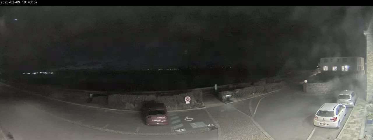 A nighttime, dimly lit parking lot with several cars, near a building and a body of water under a dark, cloudy sky.