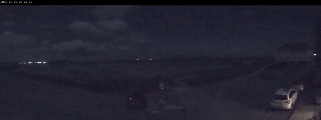 A nighttime, cloudy scene shows a dark landscape with a few distant lights, a parked white car, and other indistinct objects in a dark driveway.
