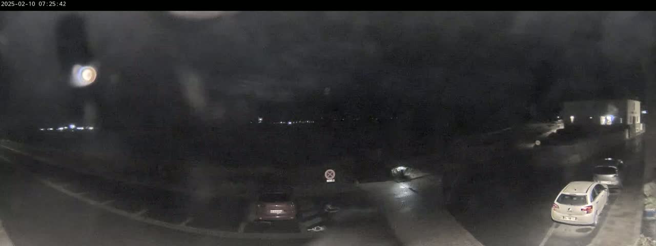 A nighttime, low-light view of a mostly empty parking lot with a few cars parked and a building in the background under a dark, cloudy sky.