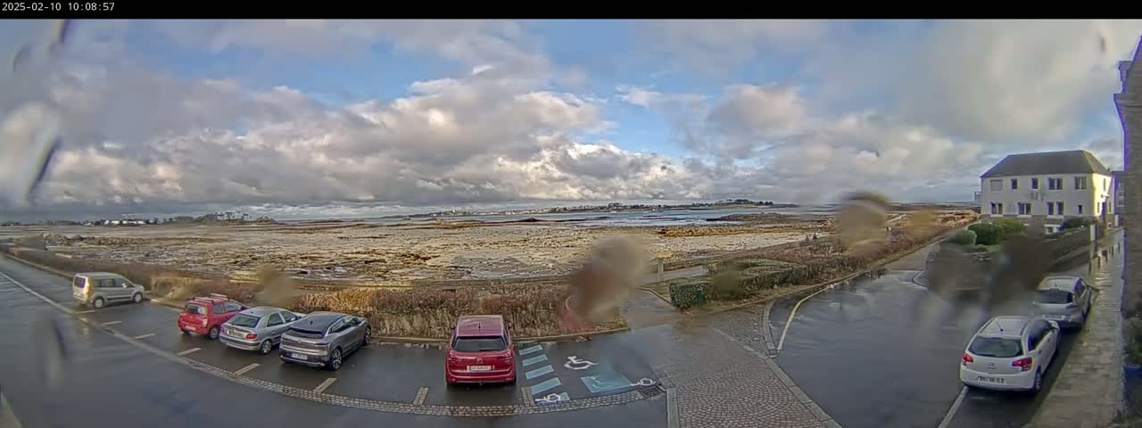 A wet parking lot with several cars is situated near a low-lying coastal area under a cloudy sky.
