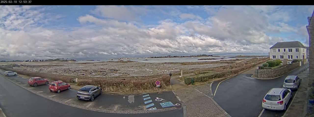 A coastal parking lot with several cars is next to a road that leads to a body of water under a partly cloudy sky.